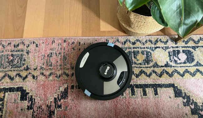 Shark robot vacuum cleaning patterned rug with hardwood floor and potted plant in peripheral